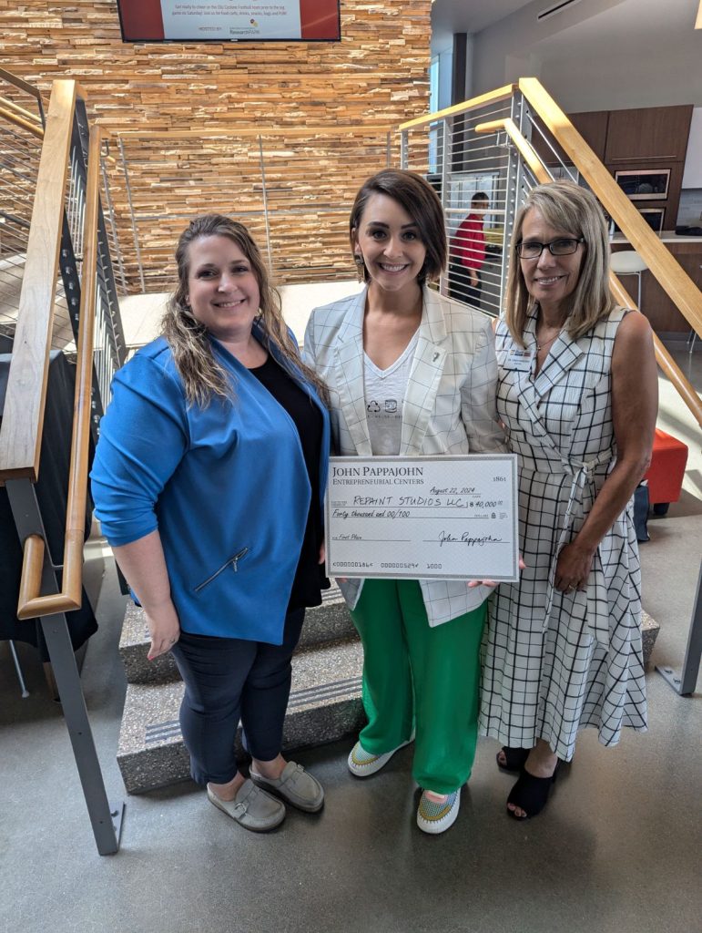 Three white women in business casual attire standing indoors. The woman in the center is holding a large check made out to Repaint Studios for the First Place award of $40,000.