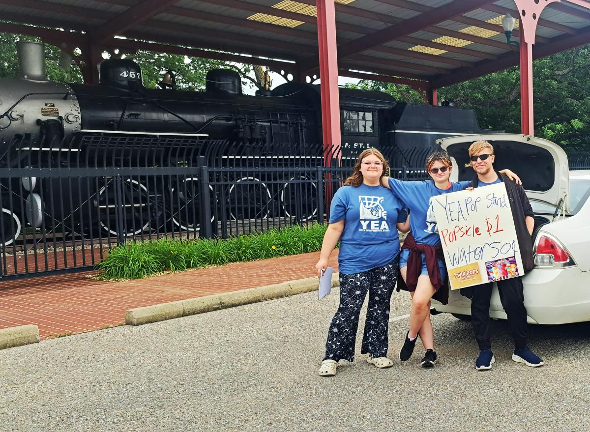 Three students wearing Youth Entrepreneurial Academy t-shirts hold a homemade sign advertising water and popsicles for sale