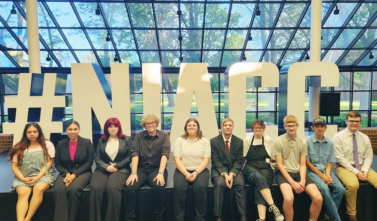 10 high school students sit on the edge of a stage in front of large metal letters that spell out "NIACC"