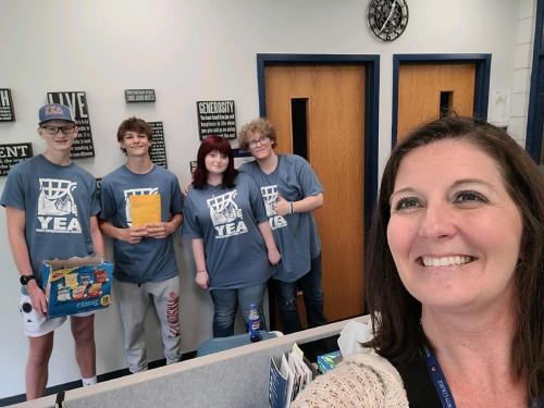 A woman takes a selfie with four high school students in the background. The students are wearing Youth Entrepreneurial Academy t-shirts