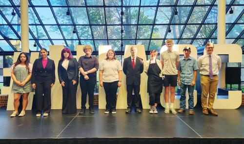 10 high school students stand on a stage in front of large metal letters that spell out "NIACC"