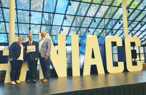 A male high school student holds a large certificate while standing next to a woman and a man in business attire