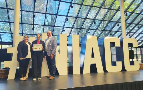 A female high school student holds a large certificate while standing next to a woman and a man in business attire